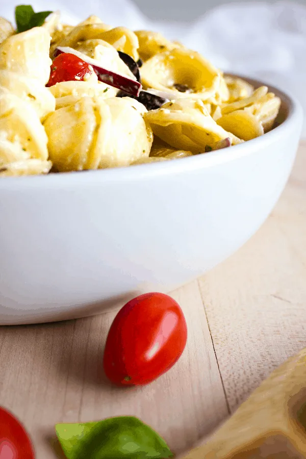 Close up of white bowl with creamy pesto pasta salad on wood surface with a grape tomato.