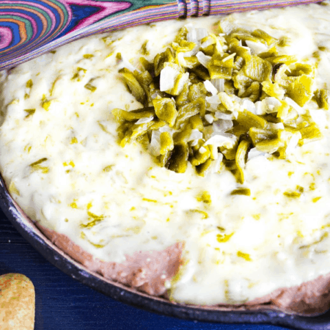 Hatch Chile Queso Bean Dip in a cast-iron skillet with a tie-dye wooden spoon laid across it and scattered corn chips on a blue surface.