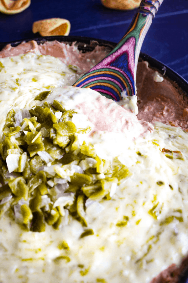 Close-up shot of hatch chile queso bean dip with a tie dye wooden spoon in it and scattered corn chips on a blue surface. 