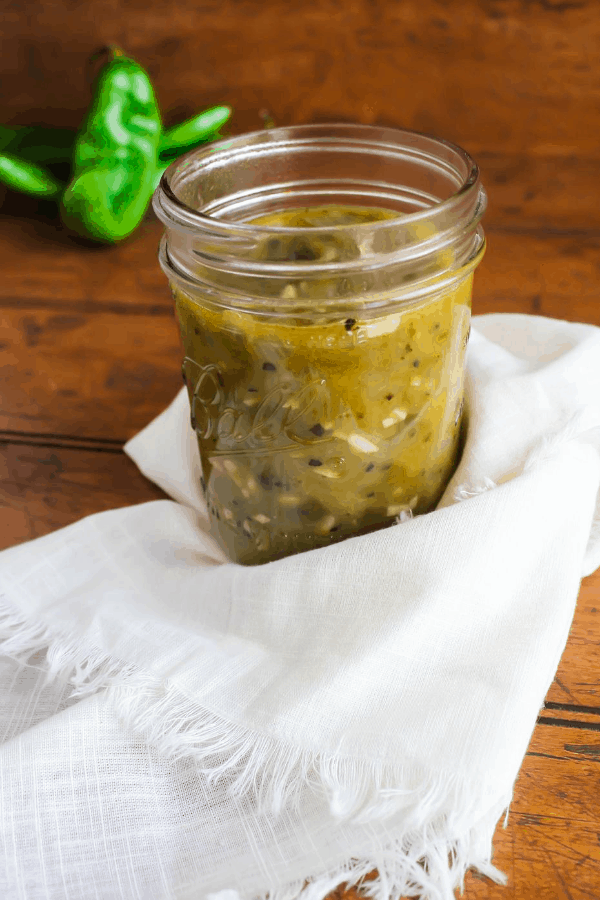 Front view of hatch green chile sauce in a clear Ball jar with a white towel and raw chiles in the background. 