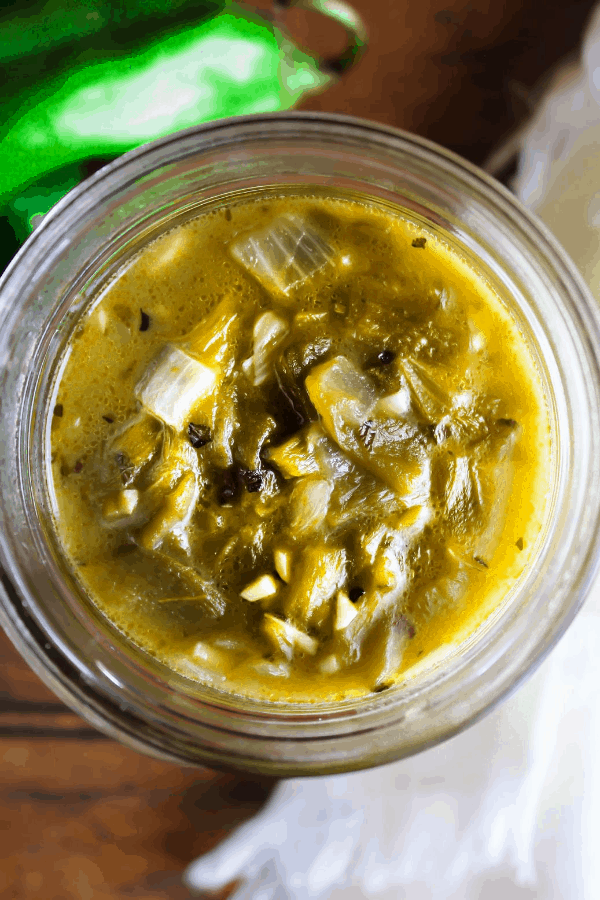 Top down shot of hatch chile sauce in a clear jar with raw chiles on a wood table.
