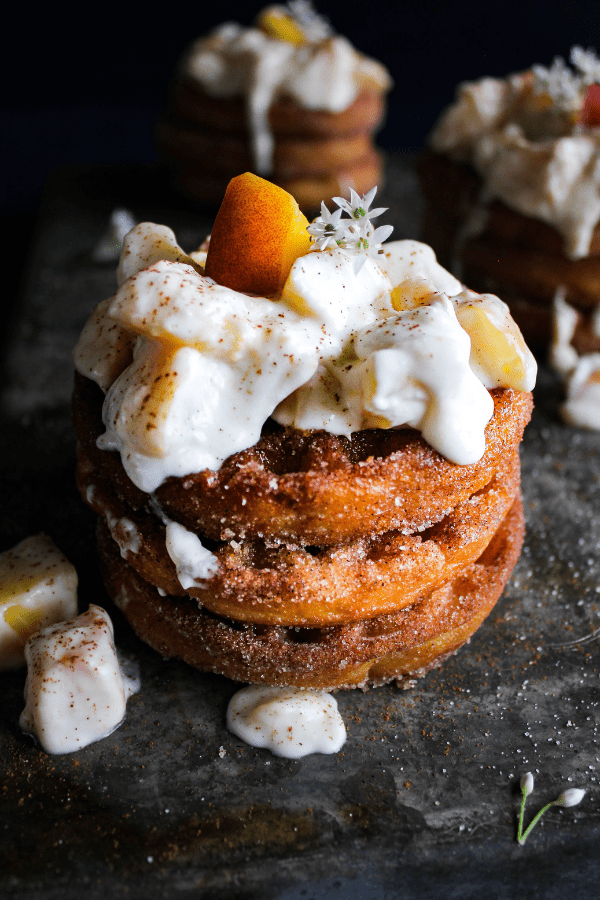 Stack of churro waffles covered in peaches an cream garnished with a fresh peach and tiny white flowers.