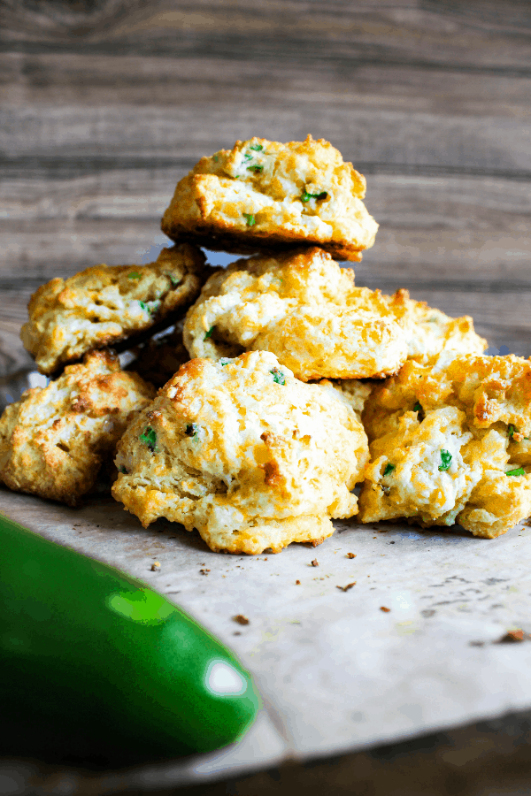 Stack of jalapeno cheddar drop biscuits on a piece of parchment paper with a jalapeno on the side. 