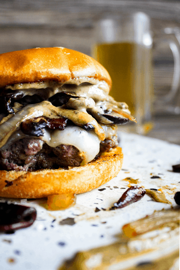 Close-up of mushroom & Swiss burger on a platter smeared with black garlic aioli with a glass mug of beer in background. 