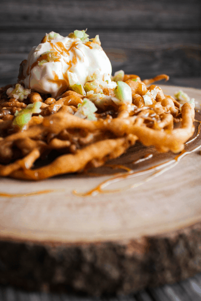 Caramel apple funnel cake topped with vanilla ice cream on a tree trunk platter.