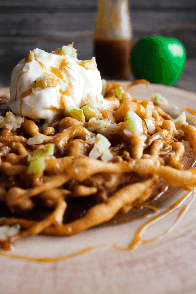 Funnel cake topped with apples, caramel, and vanilla ice cream with a green apple and bottle of caramel in background.
