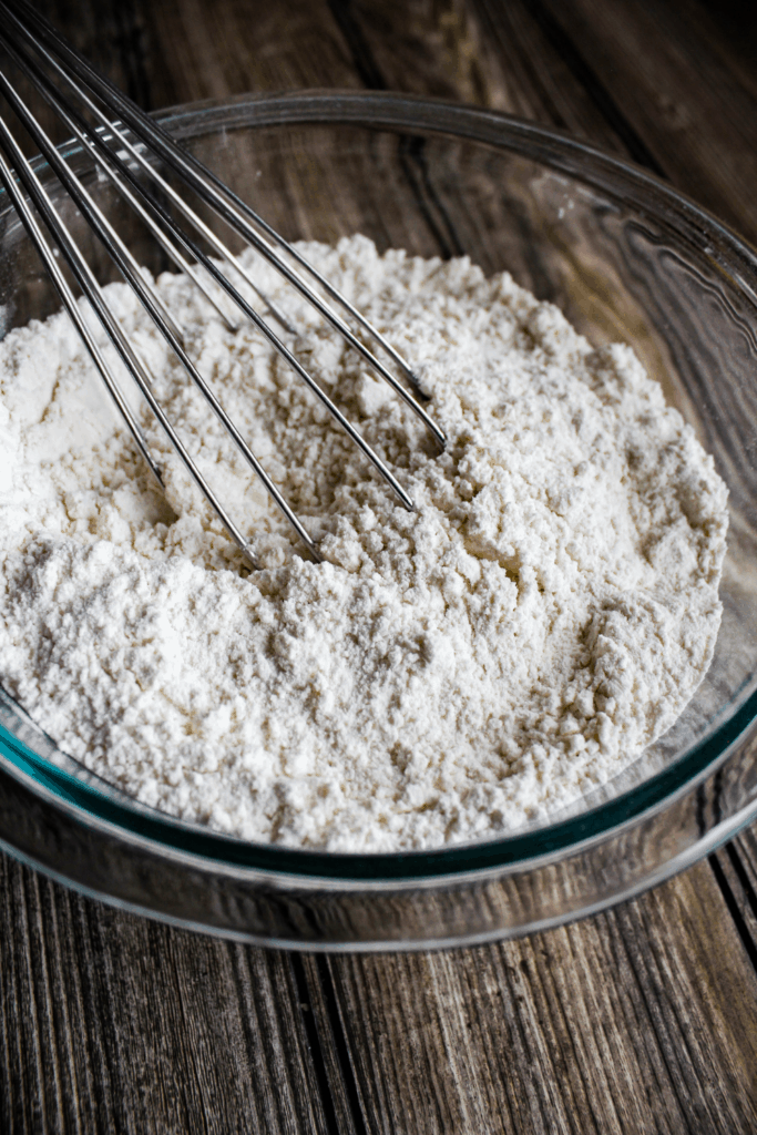 Clear glass bowl full of flour with whisk.