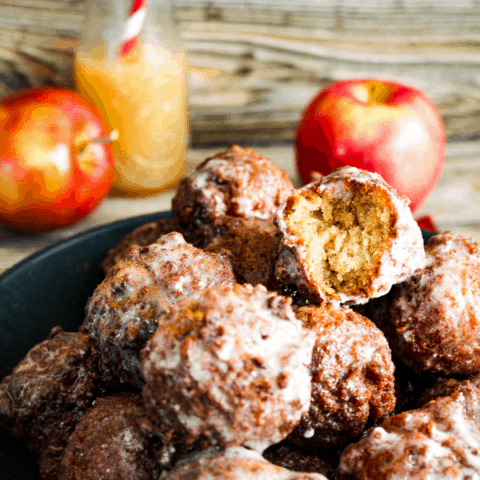 Close up of bowl of donut holes with two red apples and a jar of apple cider in background.