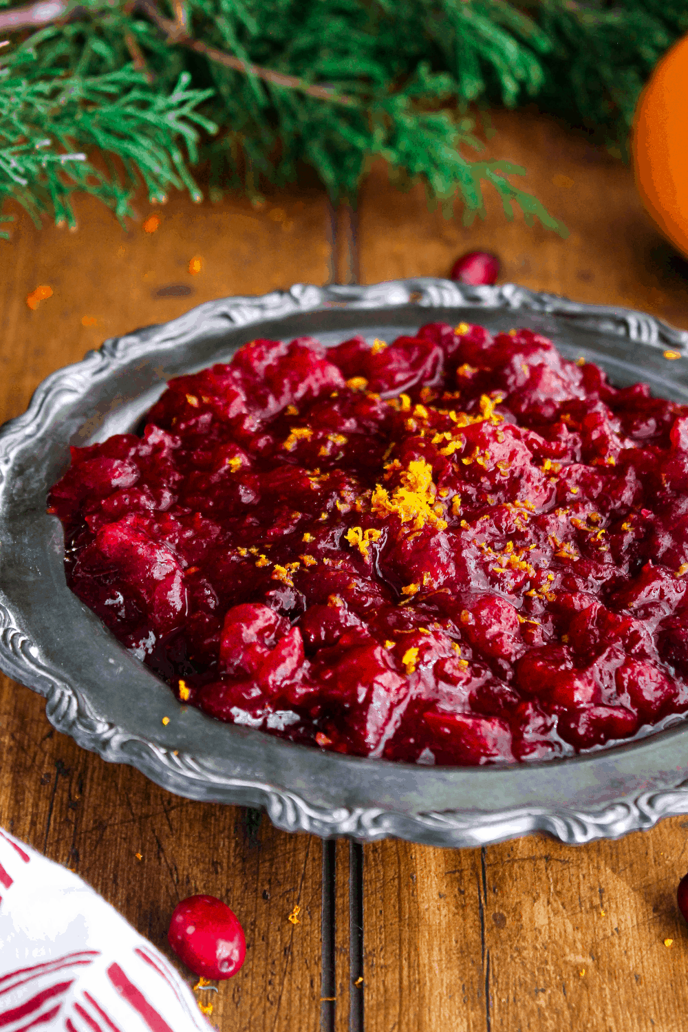 Cranberry sauce in bowl with greenery, cranberries, and an orange in the background.