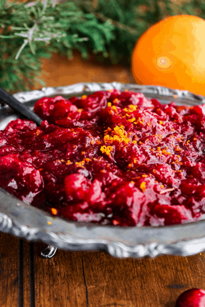 Bowl of cranberry sauce with a spoon with greenery and orange in background.