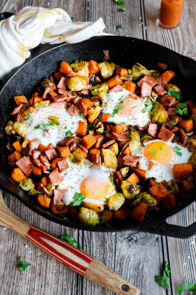 Cast iron pan filled with sweet potato Brussels sprout hash with a spatula and a dish towel.