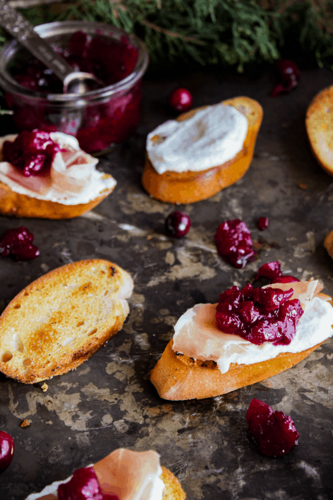 Tray of crostini in various stages of doneness with jar of cranberry sauce and greenery in background.