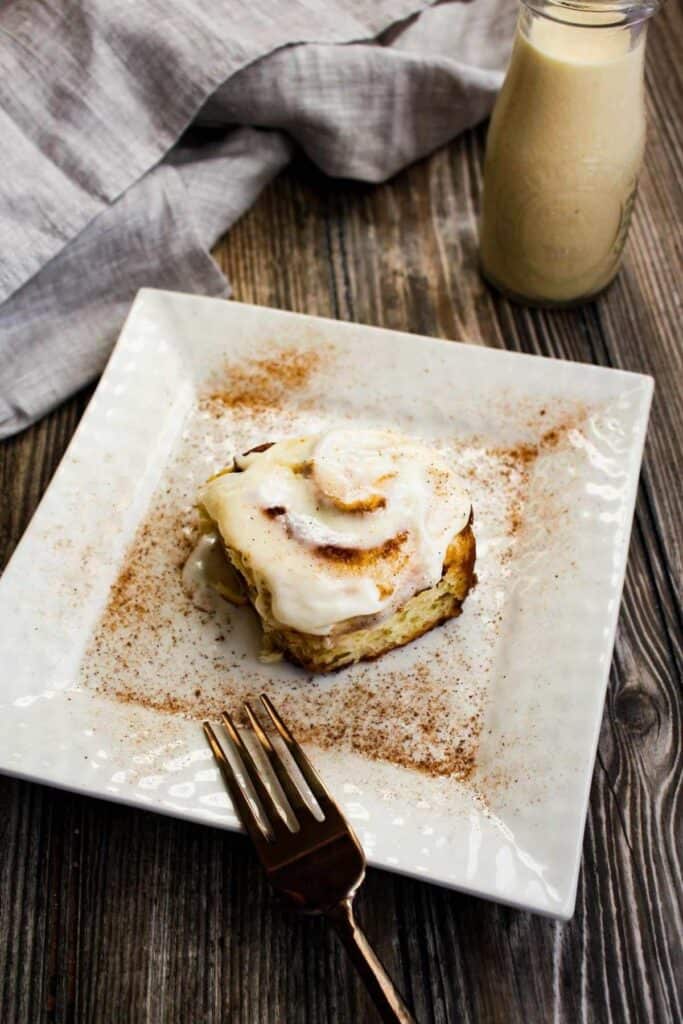Eggnog cinnamon roll on cream plate with fork and a glass of eggnog in background.