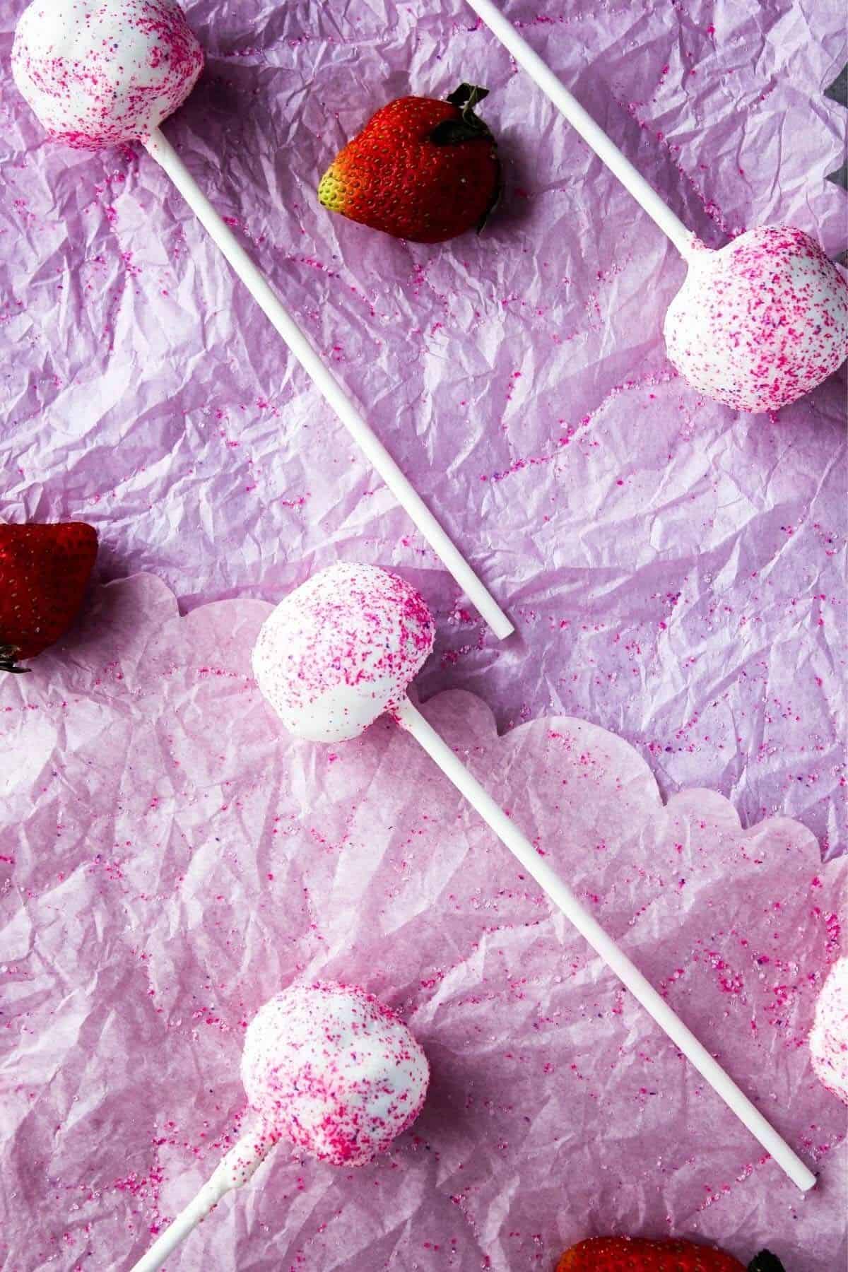 Scattered cake pops and strawberries on pink surface.