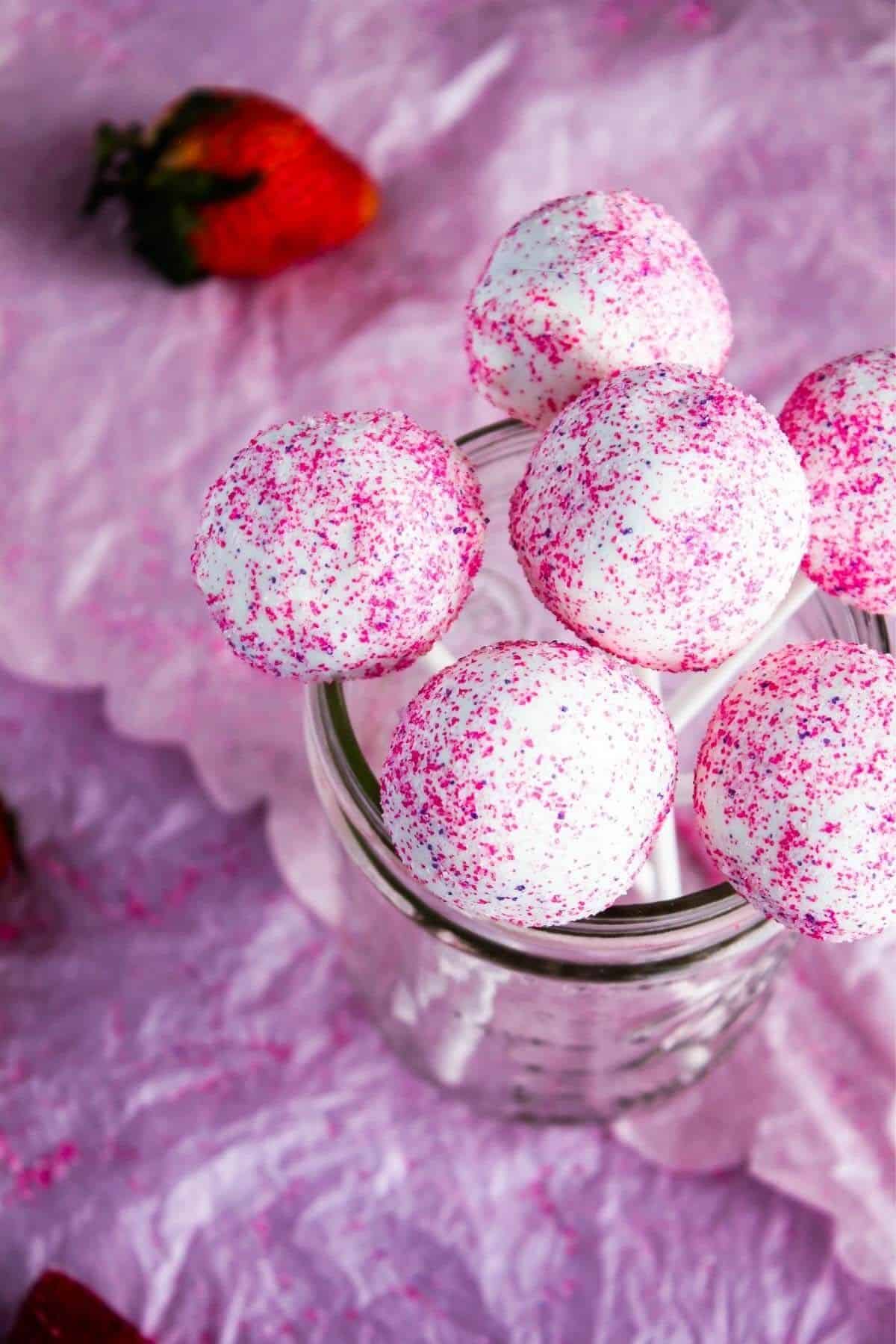 Glass jar filled with cake pops with scattered strawberries.