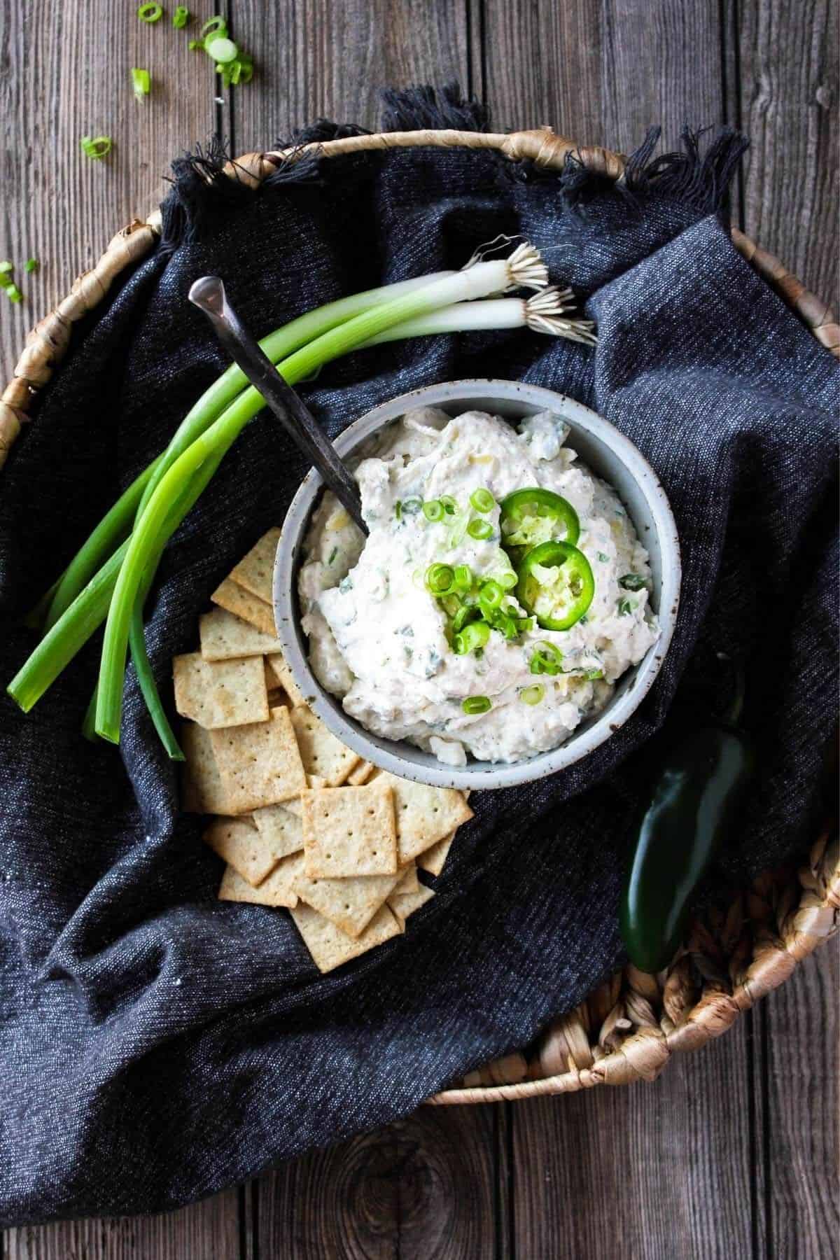 Wicker track with blue towel bowl of artichoke dip with jalapenos and crackers.