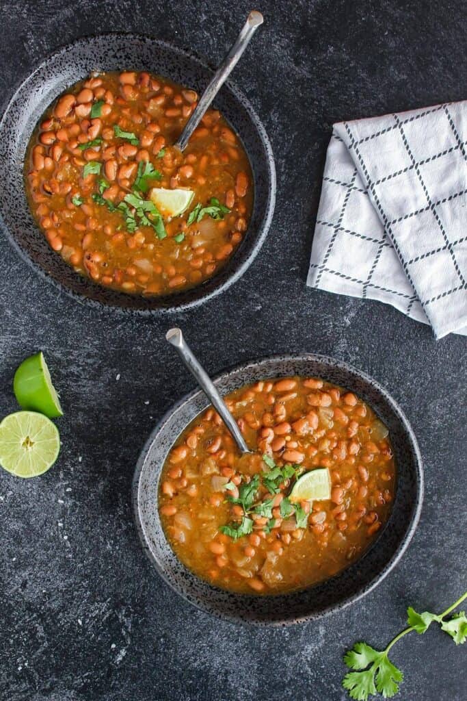 Two bowls of salsa verde pinto beans with lime halves and a towel.