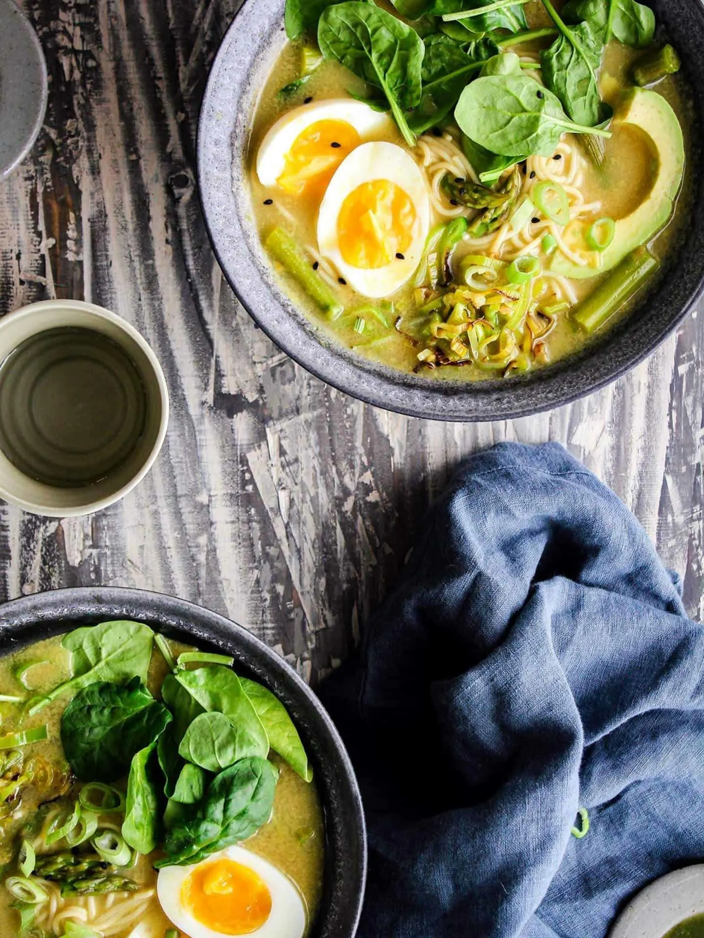 Two bowls of green goddess ramen with a blue towel and small bowls of condiments.