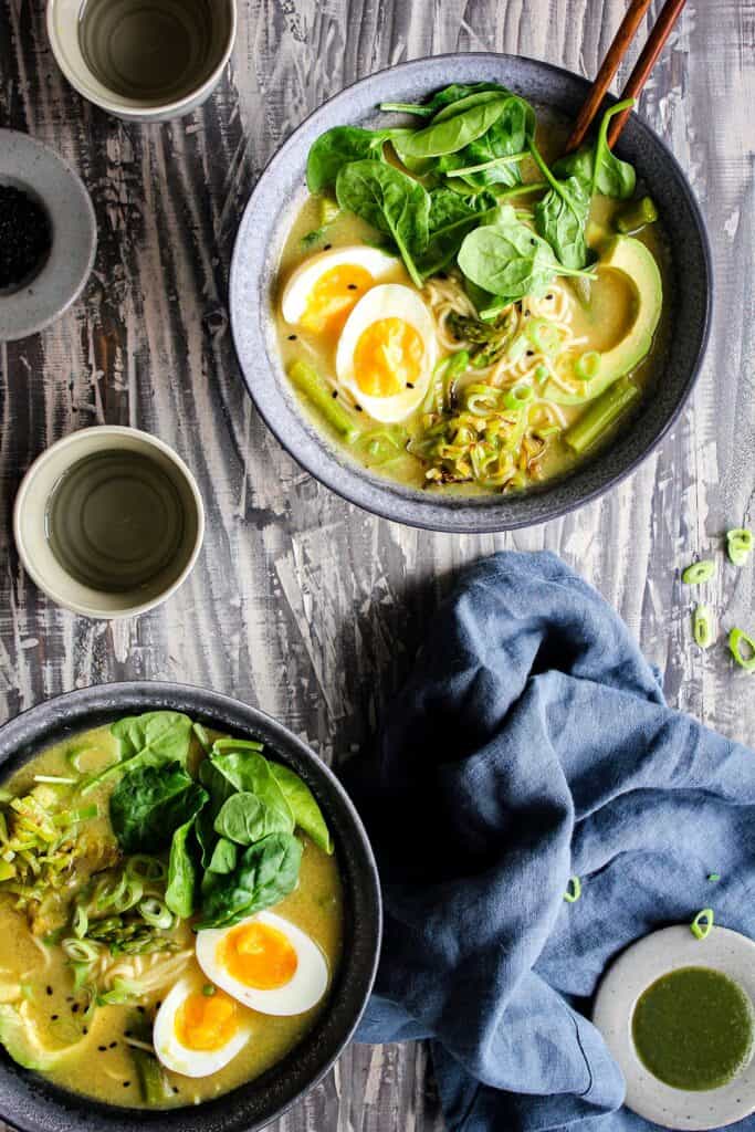Two bowls of green goddess ramen with a blue towel and small bowls of condiments.