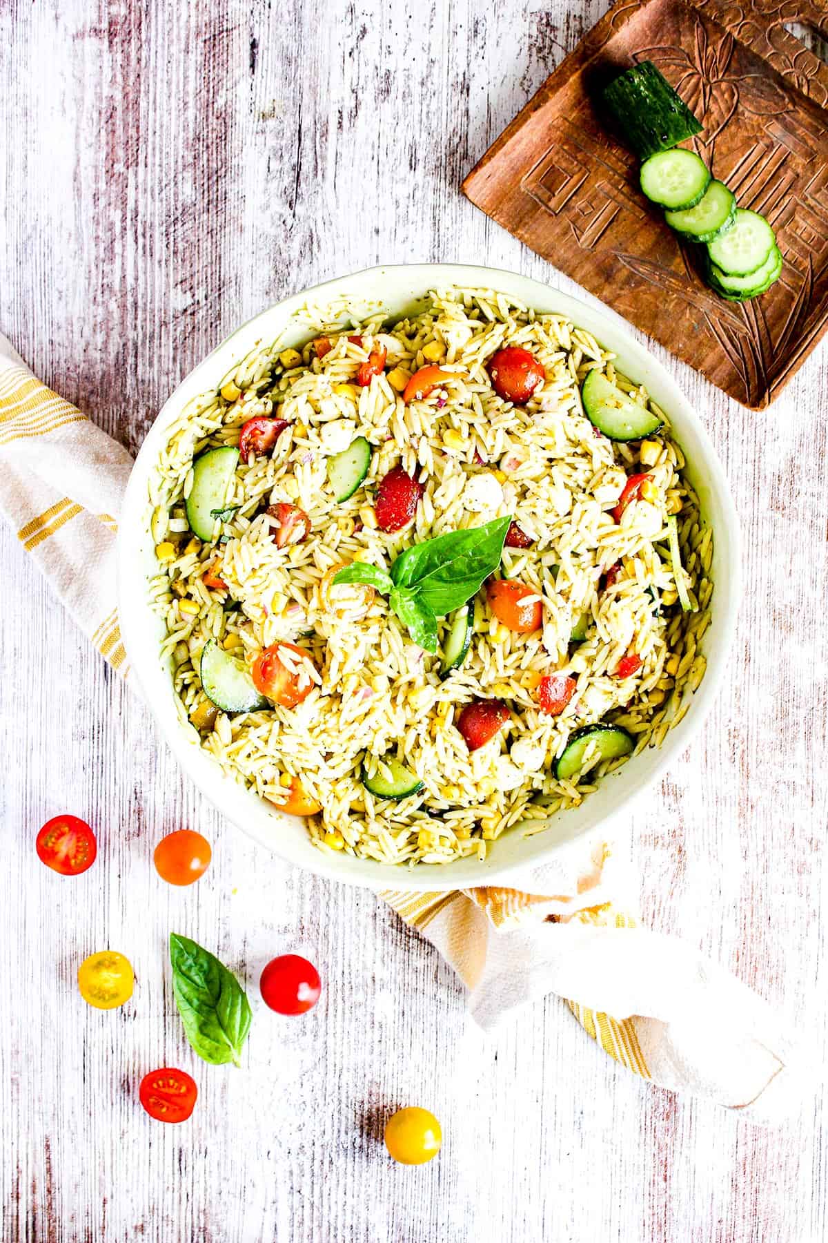 Serving bowl of orzo pesto salad with summer vegetables, a towel, tomatoes, and sliced cucumber. 