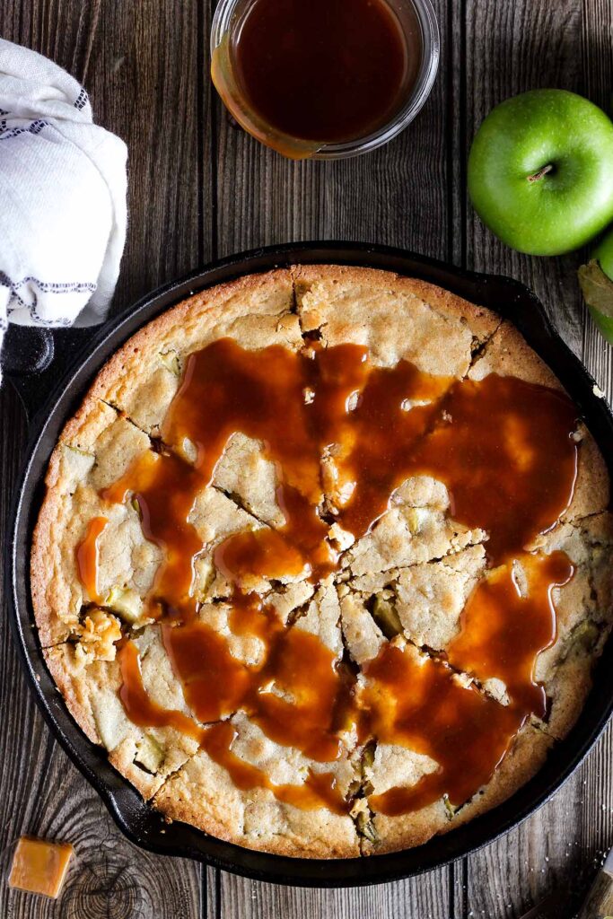 Apple blondies in a cast-iron skillet with an apple, caramels, and caramel sauce.