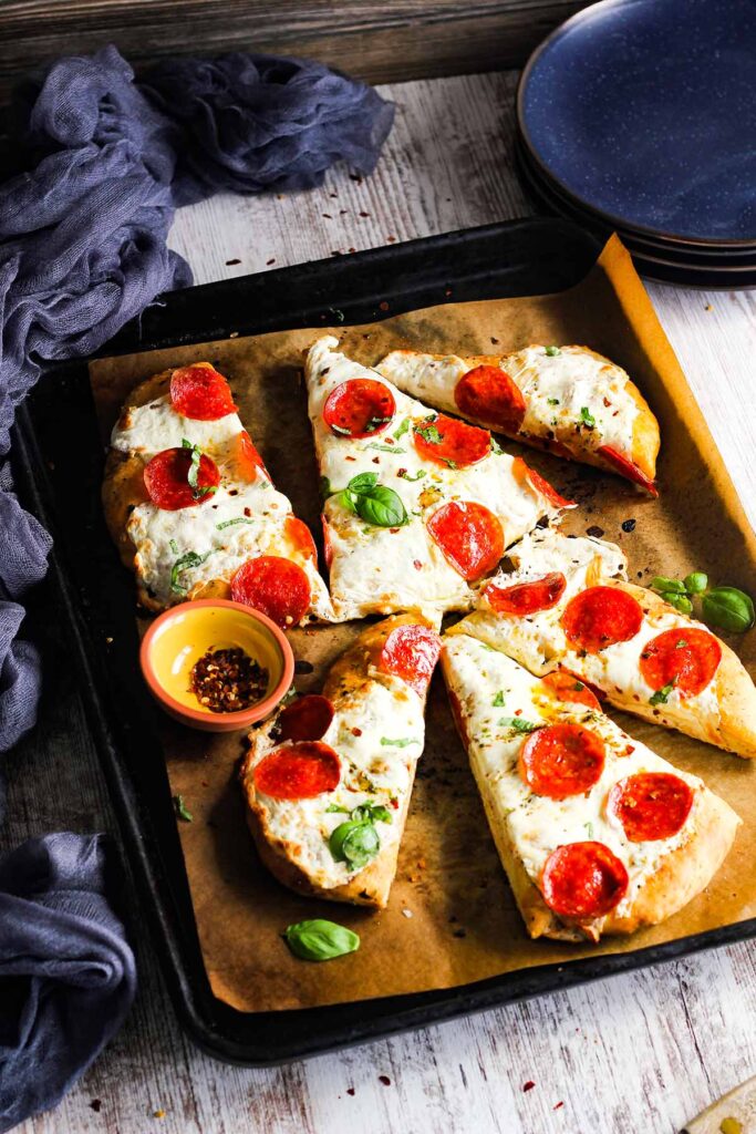 Sheet pan with sliced pepperoni flatbread and bowl of red pepper flakes.