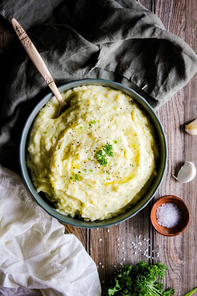 Boursin cheese mashed potatoes in a bowl surrounded by two dish towels, garlic cloves, parsley, and a bowl of salt.