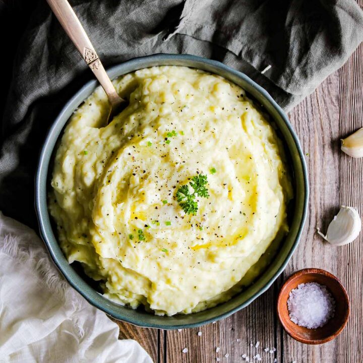 Boursin cheese mashed potatoes in a bowl surrounded by two dish towels, garlic cloves, parsley, and a bowl of salt.