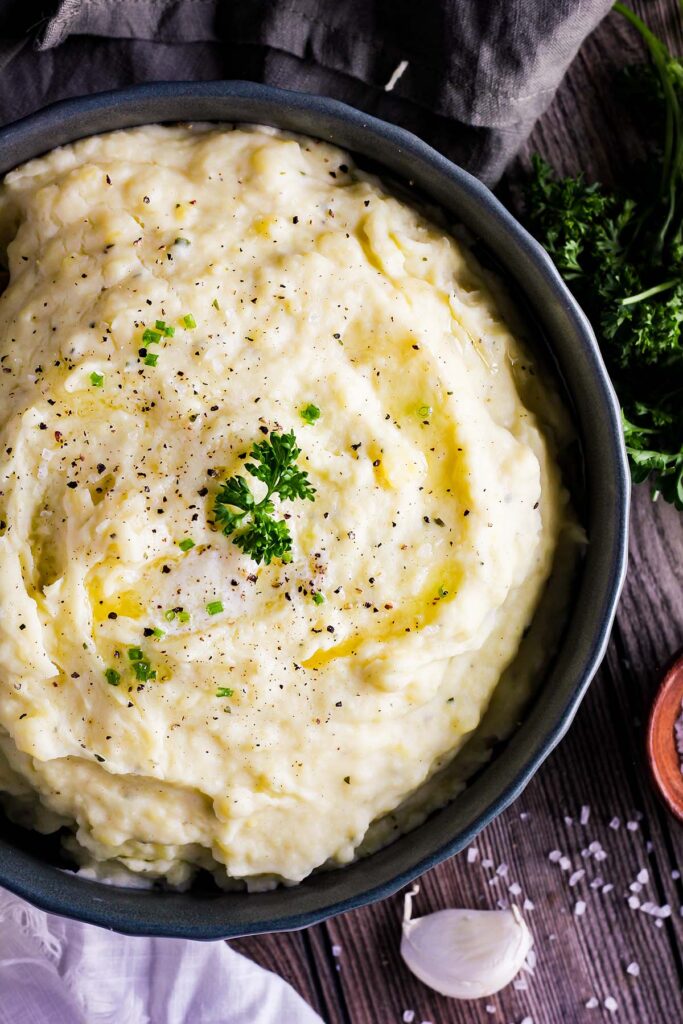 Bowl of Boursin mashed potatoes surrounded by garlic clove, salt, and parsley.
