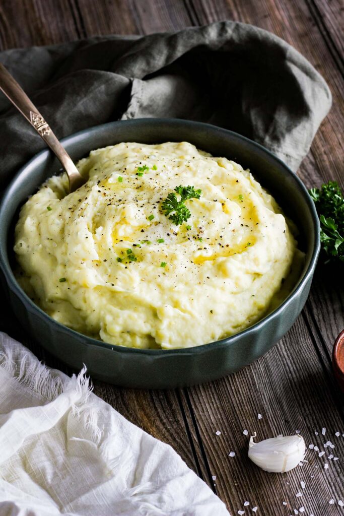 Bowl of mashed potatoes with a spoon with parsley and a clove of garlic.