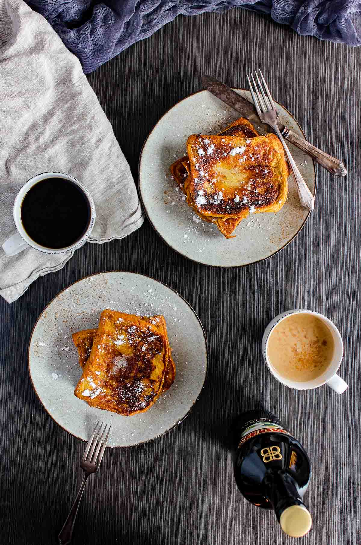Two plates of Baileys French toast with two cups of coffee and a bottle of Baileys Irish Cream.