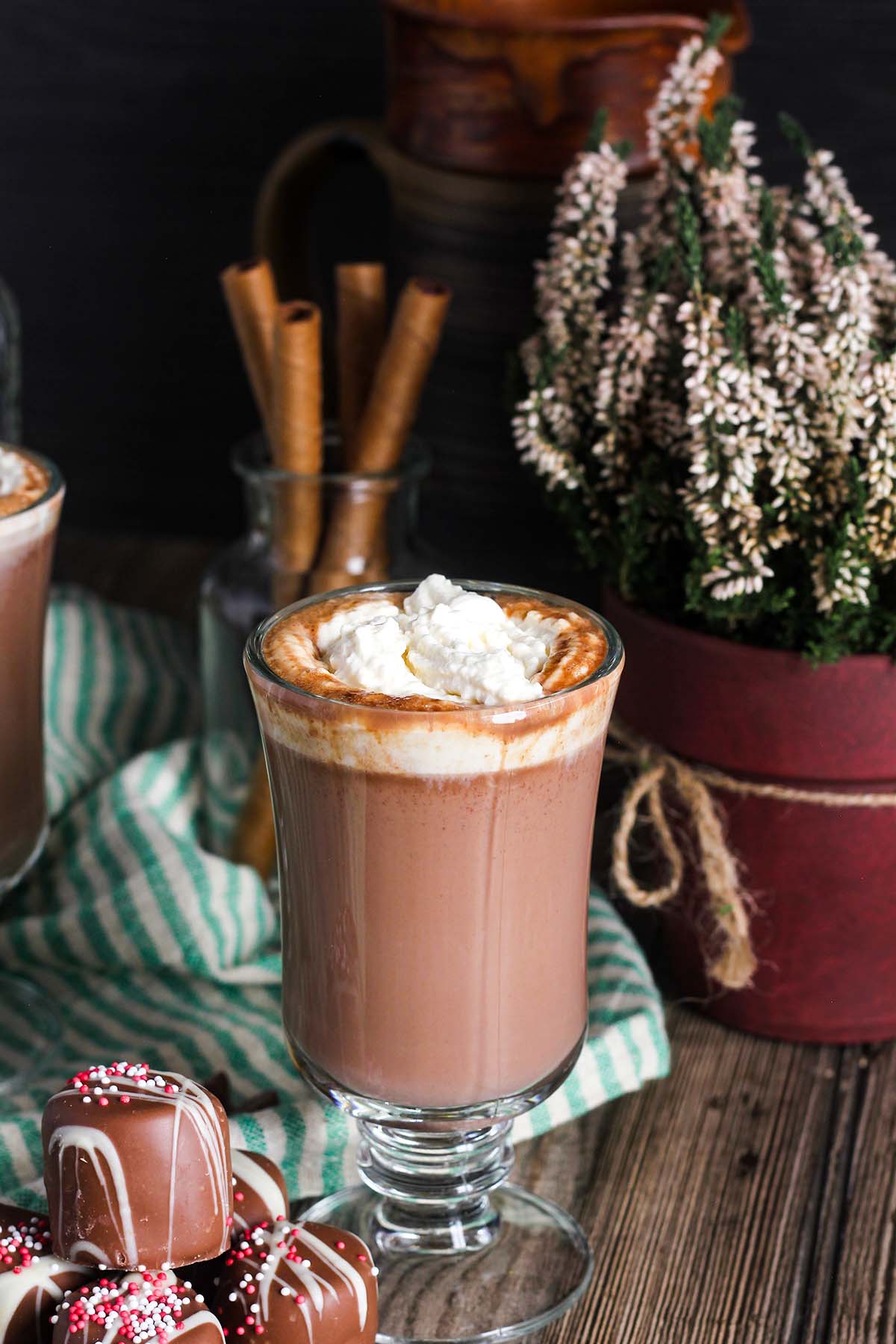 A glass of Amaretto hot chocolate next to a stack of chocolate covered marshmallows with a plant and a pitcher in the background.
