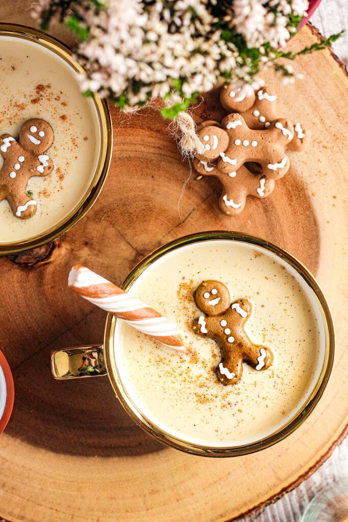 Two glasses of Baileys in eggnog garnished with gingerbread man and candy cane next to stack of gingerbread men and a plant.