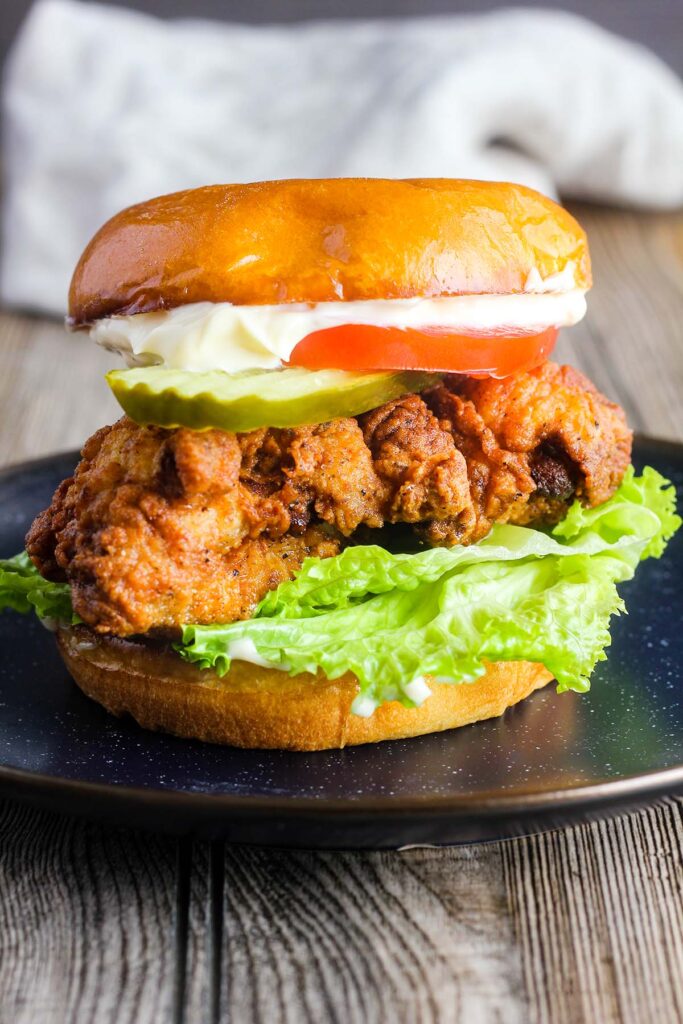 A crispy chicken sandwich with lettuce, tomato, pickle, and mayo on a blue plate with a dish towel in the background.