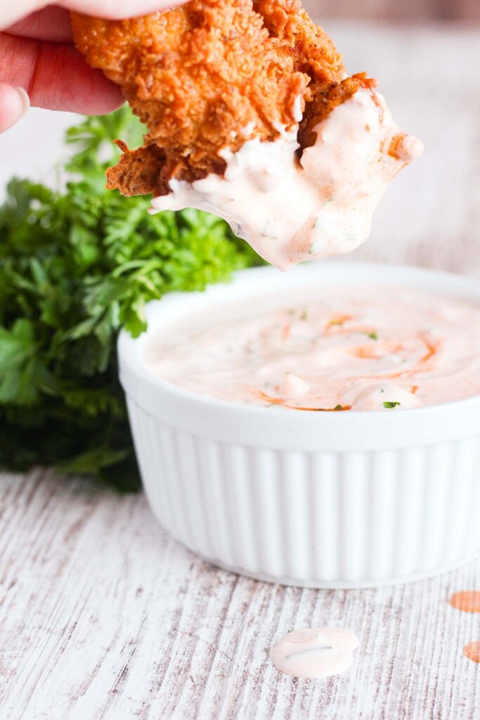 A hand holding a chicken tender dipped in buffalo ranch above a bowl of ranch.