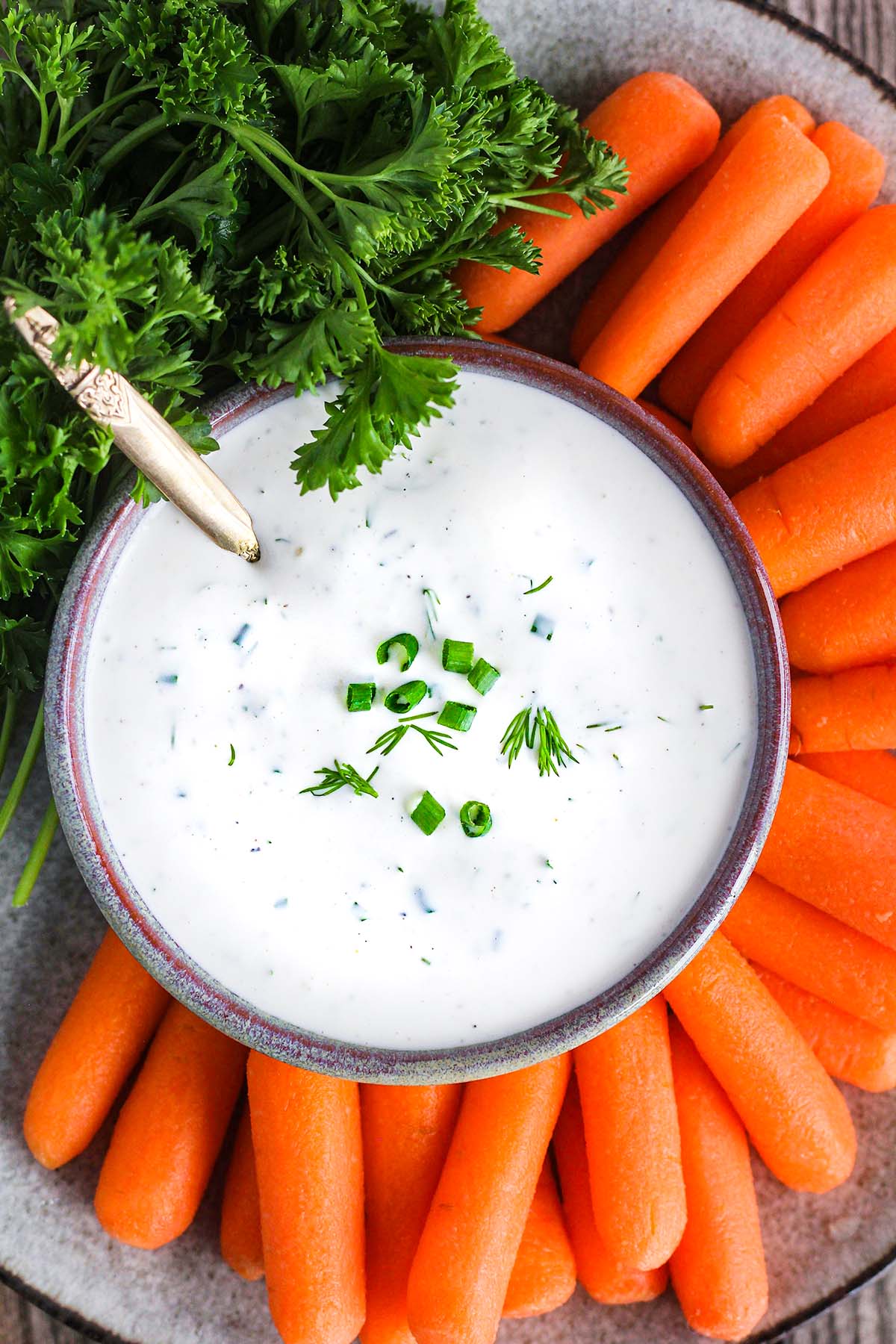 Bowl of garlic ranch dressing with a spoon surrounded by baby carrots and bunch of parsley