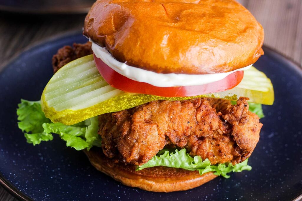 Buttermilk chicken sandwich dressed with lettuce, tomato, pickle, and may on a dark blue plate.