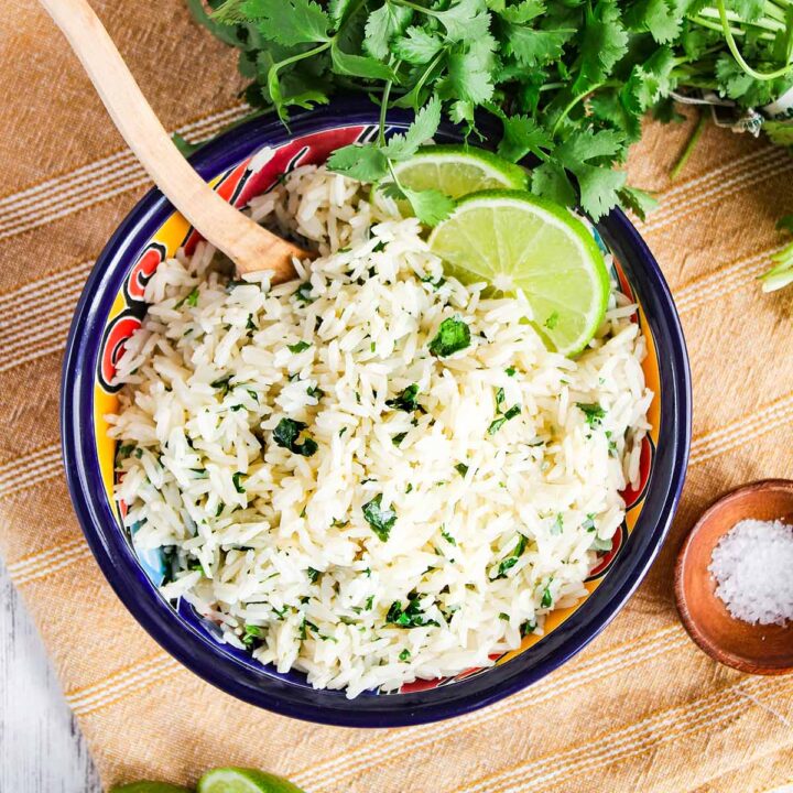 Cilantro rice in a bowl garnished with lime slices surrounded by bunch of cilantro, lime halves, and small bowl of salt.