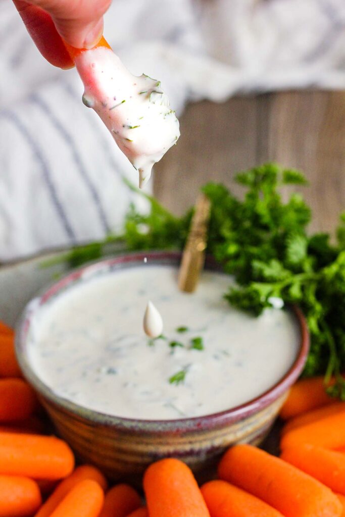 Fingers holding a carrot dripping with ranch with bowl of ranch and carrots in the background