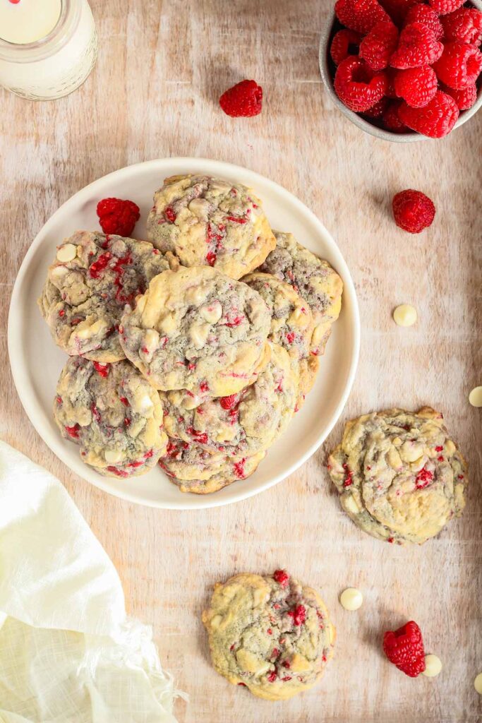 Stack of raspberry white chocolate chip cookies on a plate surrounded by a glass of milk, scattered cookies, and a bowl of raspberries