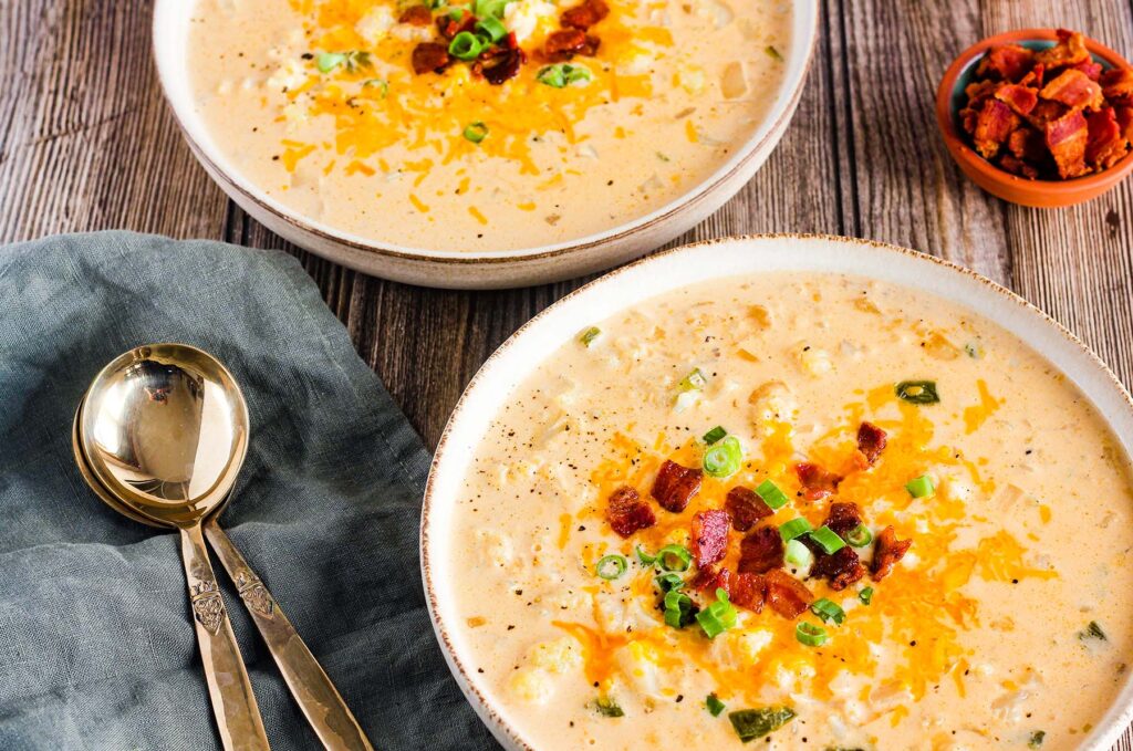 Two bowls of jalapeno popper cauliflower soup next to two spoons and a small bowl of bacon bits.