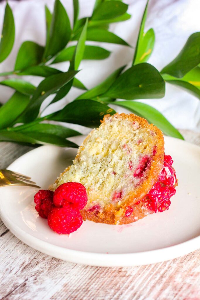 A single slice of white chocoalte bundt cake with raspberries on a white plate with greenery in background.