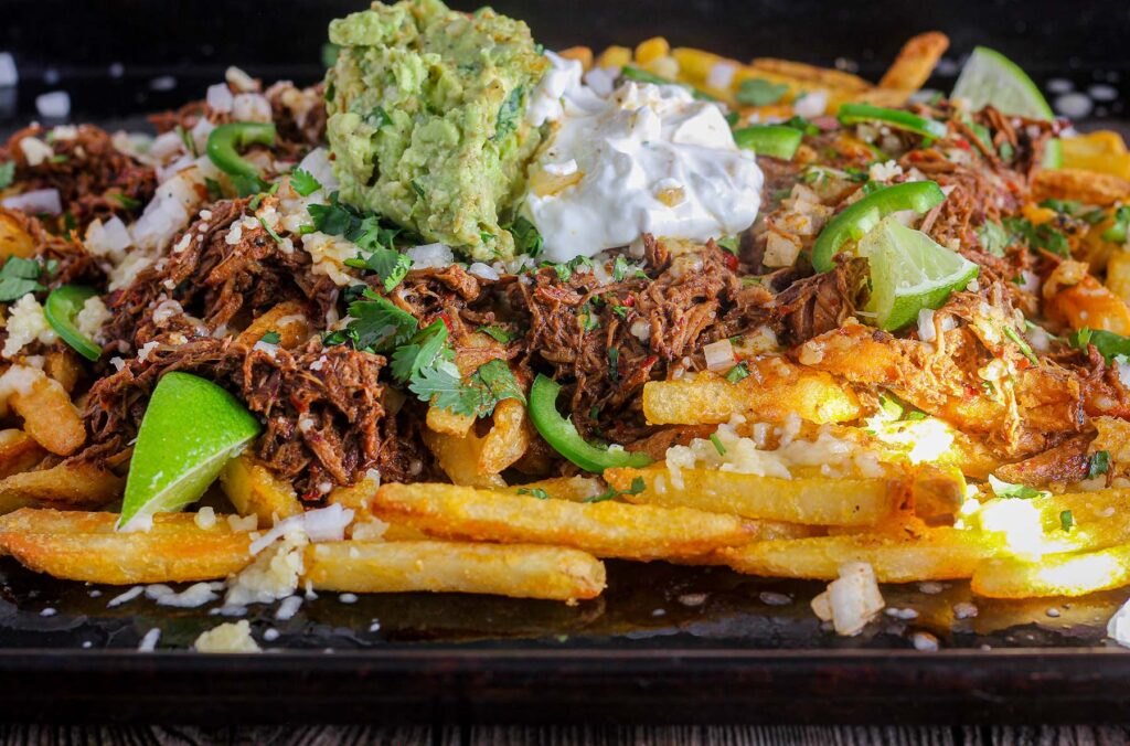 Birria fries on a sheet pan topped with lime wedges, sliced jalapenos, guacamole, and sour cream.