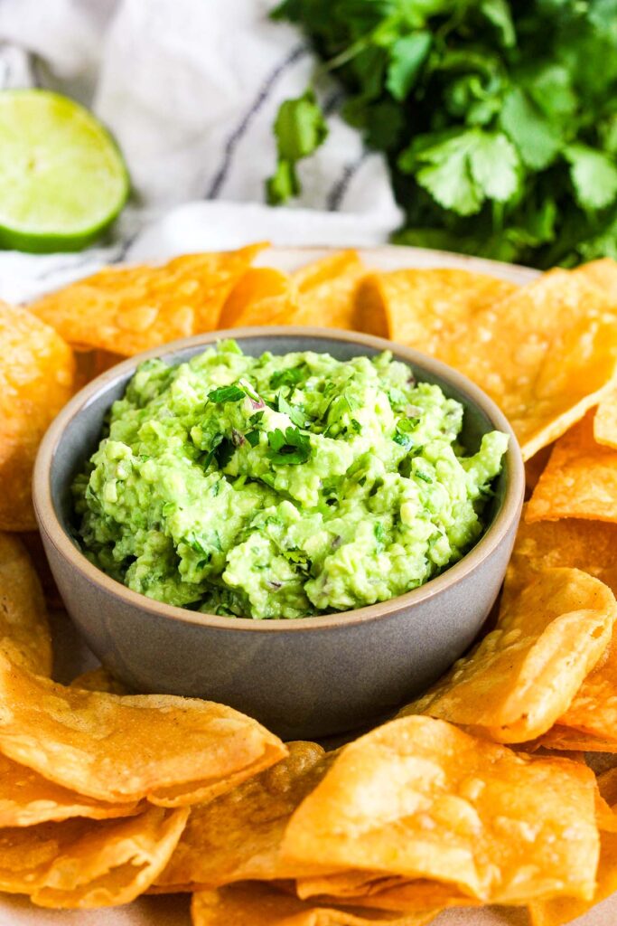Bowl of guacamole surrounded by tortilla chips with a lime and cilantro in the background.