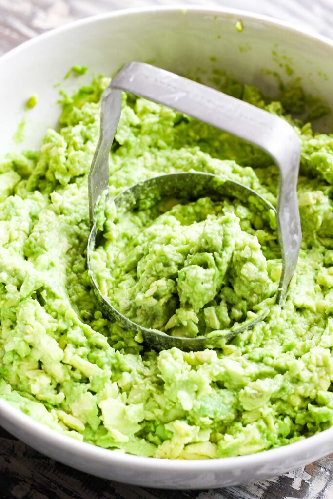 Bowl of mashed avocado with a manual food chopper in the center.