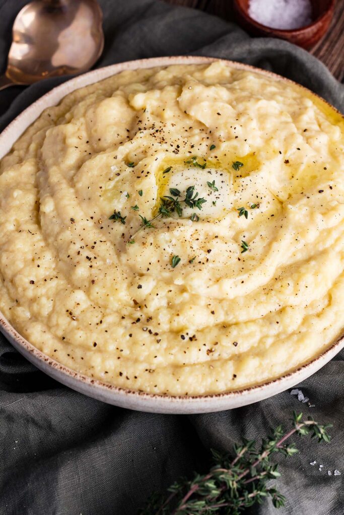 Bowl of mashed miso potatoes garnished with butter and thyme with a spoon and bowl of salt.