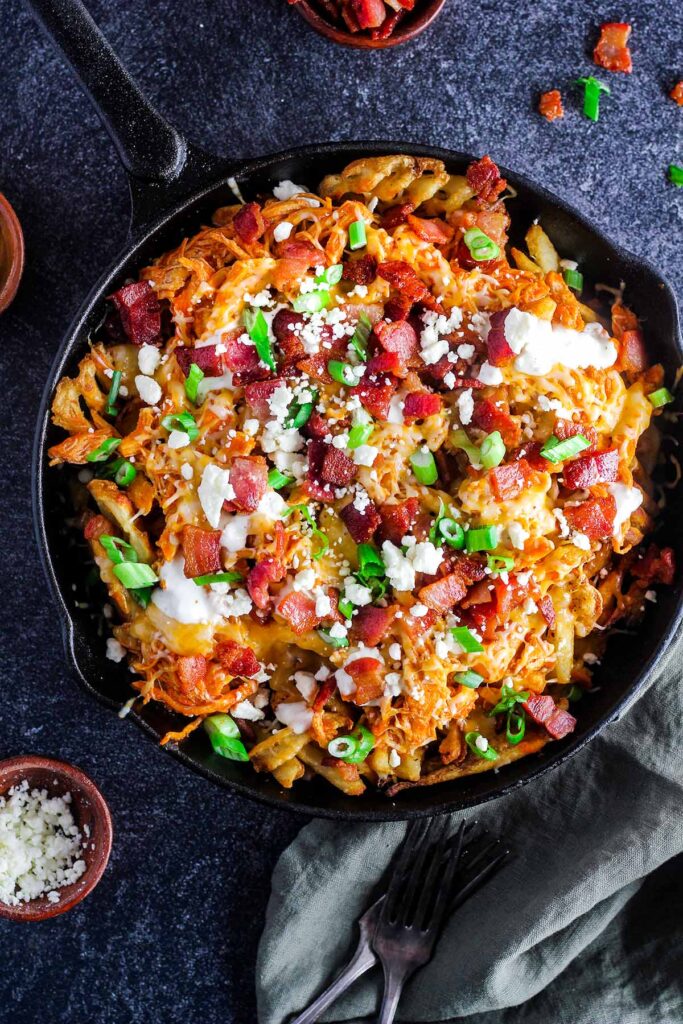 Buffalo chicken fries in a cast-iron skillet with a small bowl of bacon bits and cheese.