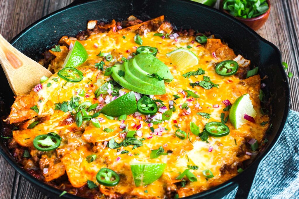 Cast-iron skillet enchiladas topped with lime, cilantro, onion, and avocado with a wooden spoon.