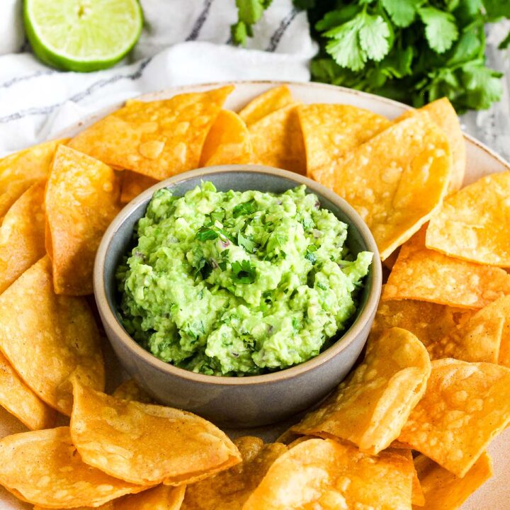 Plate of tortilla chips with a bowl of guacamole in the center and a lime and bunch of cilantro in background.