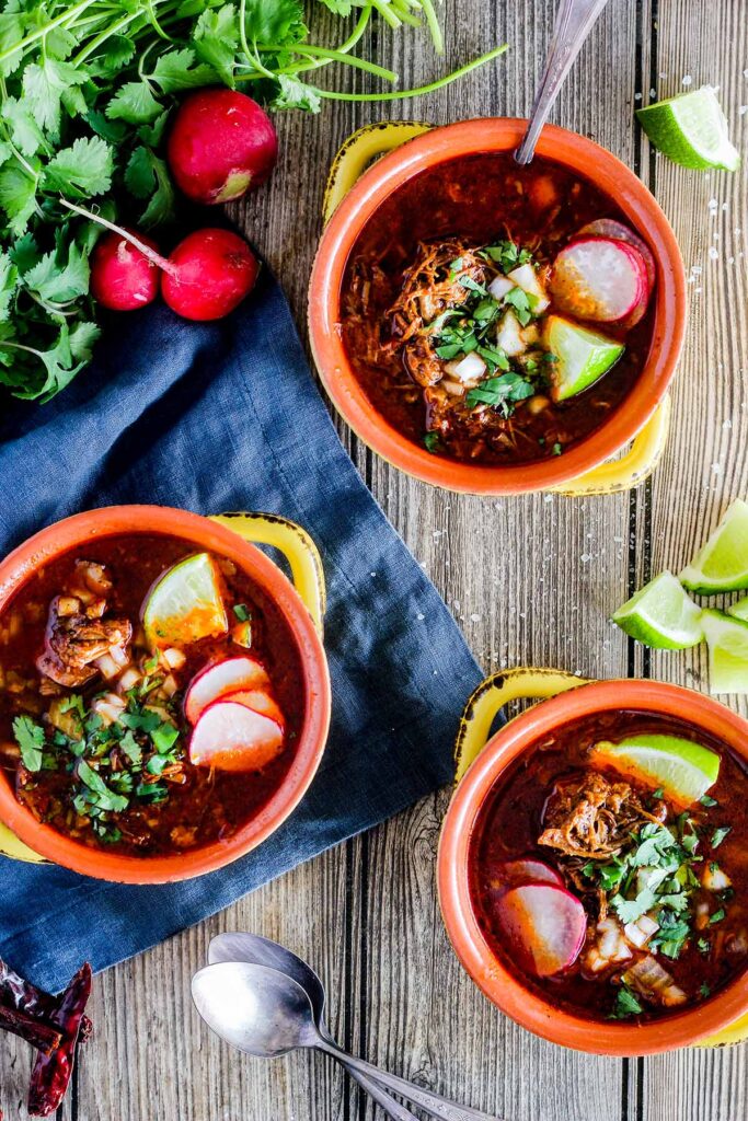 Three garnished bowls of beef birria stew surrounded by lime wedges, cilantro, radishes, and two spoons.